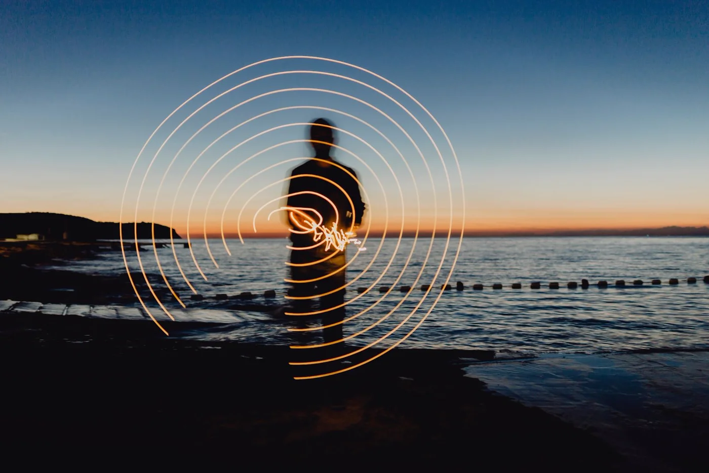 A silhouetted figure at twilight with spiralling rings of golden light drawn around them by the sea